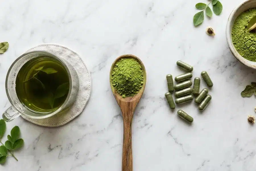 Différentes formes de consommation du moringa : poudre, gélules et infusion Poudre de moringa dans une cuillère, gélules de moringa et verre de tisane verte sur surface en marbre