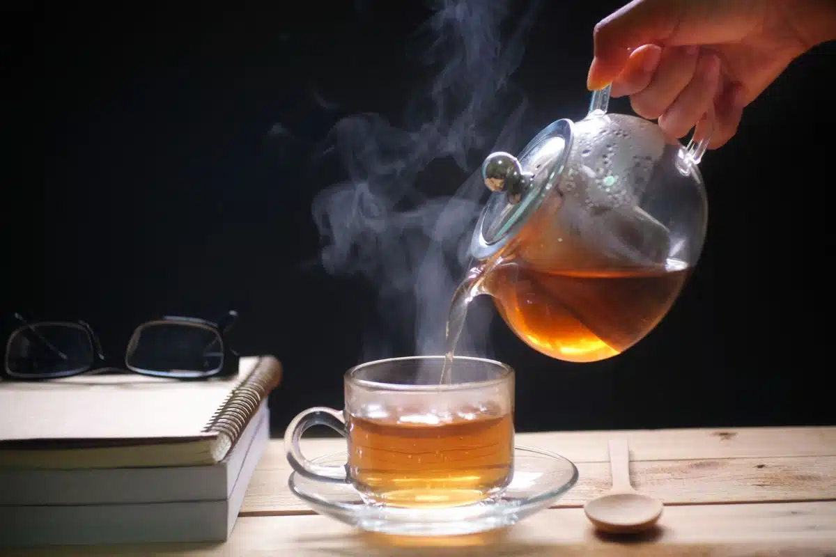 Thé chaud versé d'une théière en verre dans une tasse fumante, à côté de livres