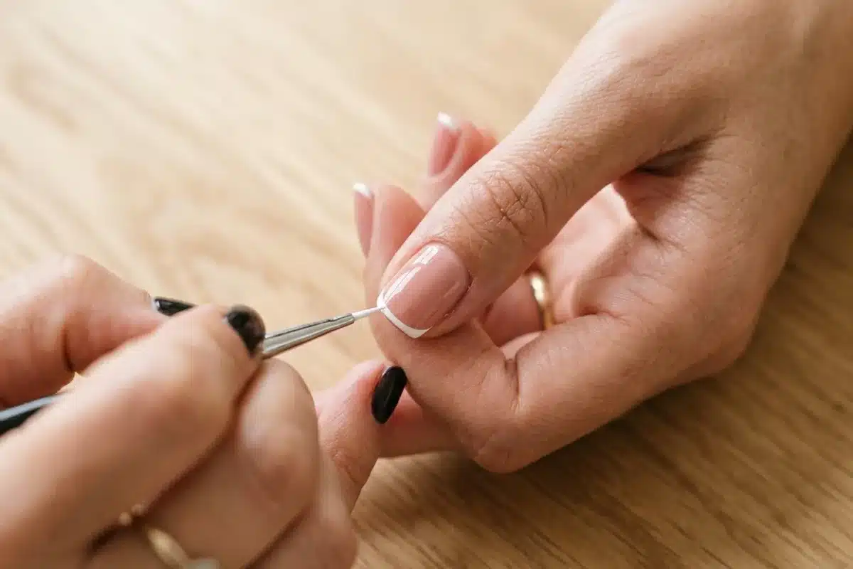 Application french manucure fine Gros plan sur des ongles naturels avec l'application d'une french fine, trait blanc ultra-mince sur fond bois clair